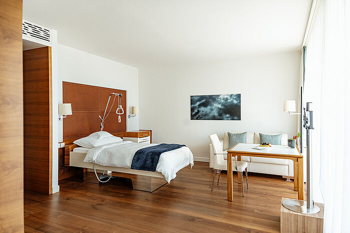 Helles, modernes Zimmer mit Holzboden, einem Bett, einem Sofa und einem Schreibtisch mit rosa Blumen. Ein großes Fenster öffnet sich zu einem malerischen Balkonblick. Ruhiges Ambiente.