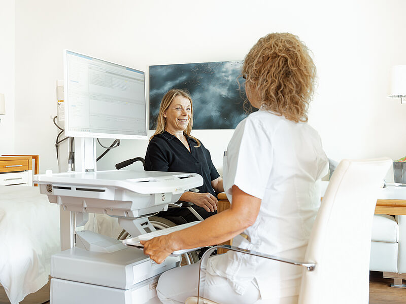 Eine ältere Frau entspannt sich auf einem Liegestuhl und lächelt eine Krankenschwester an, die neben ihr sitzt und ein Klemmbrett hält. In der Nähe steht ein Tisch mit Frühstücksartikeln, der eine ruhige, fürsorgliche Atmosphäre vermittelt.