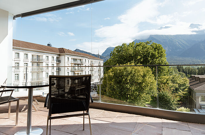 Sonniger Balkon mit einem kleinen Tisch für das Frühstück mit Saft und Gebäck. Mit Blick auf üppige Bäume und majestätische Berge unter einem klaren Himmel.