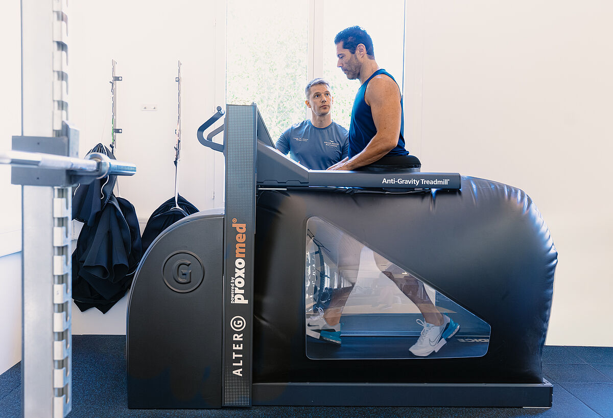 Eine ältere Frau trainiert auf einem Anti-Schwerkraft-Laufband, angeleitet von einem jungen männlichen Trainer. Der helle, geräumige Raum strahlt eine unterstützende, energiegeladene Atmosphäre aus.